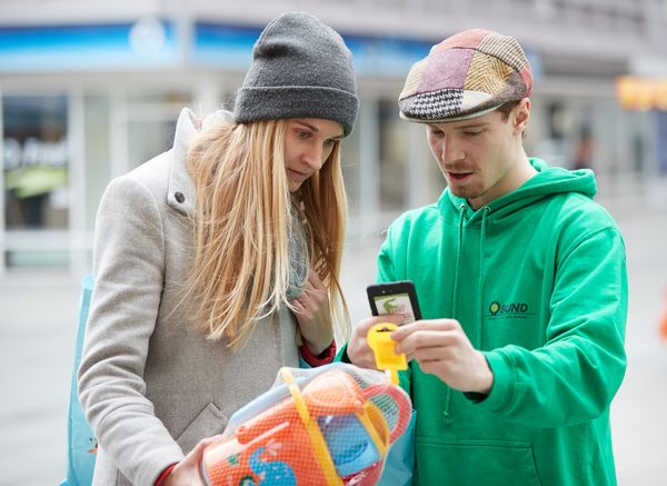 Aktivist des BUND scannt mit dem Smartphone einen Spielwarenartikel, den eine Verbraucherin ihm präsentiert.
